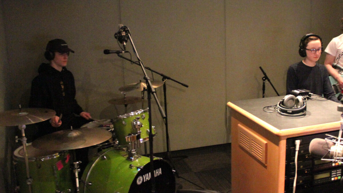 Two teens wearing headphones performing on piano keyboard and drum set in a radio studio