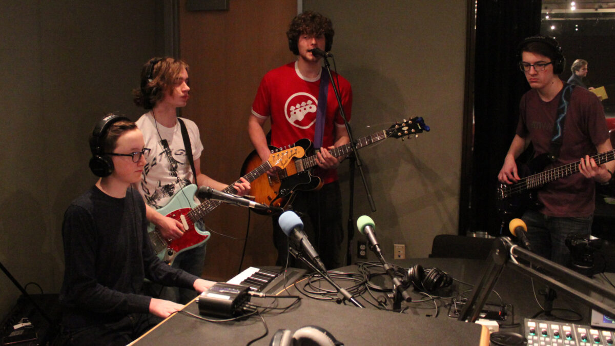 Four teens wearing headphones performing on piano keyboard, two guitars, and bass in a radio studio, one of the guitarists singing into a microphone