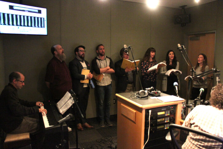 An adult playing a piano keyboard and looking at sheet music on a music stand and seven adults singing and holding scripts in a radio studio