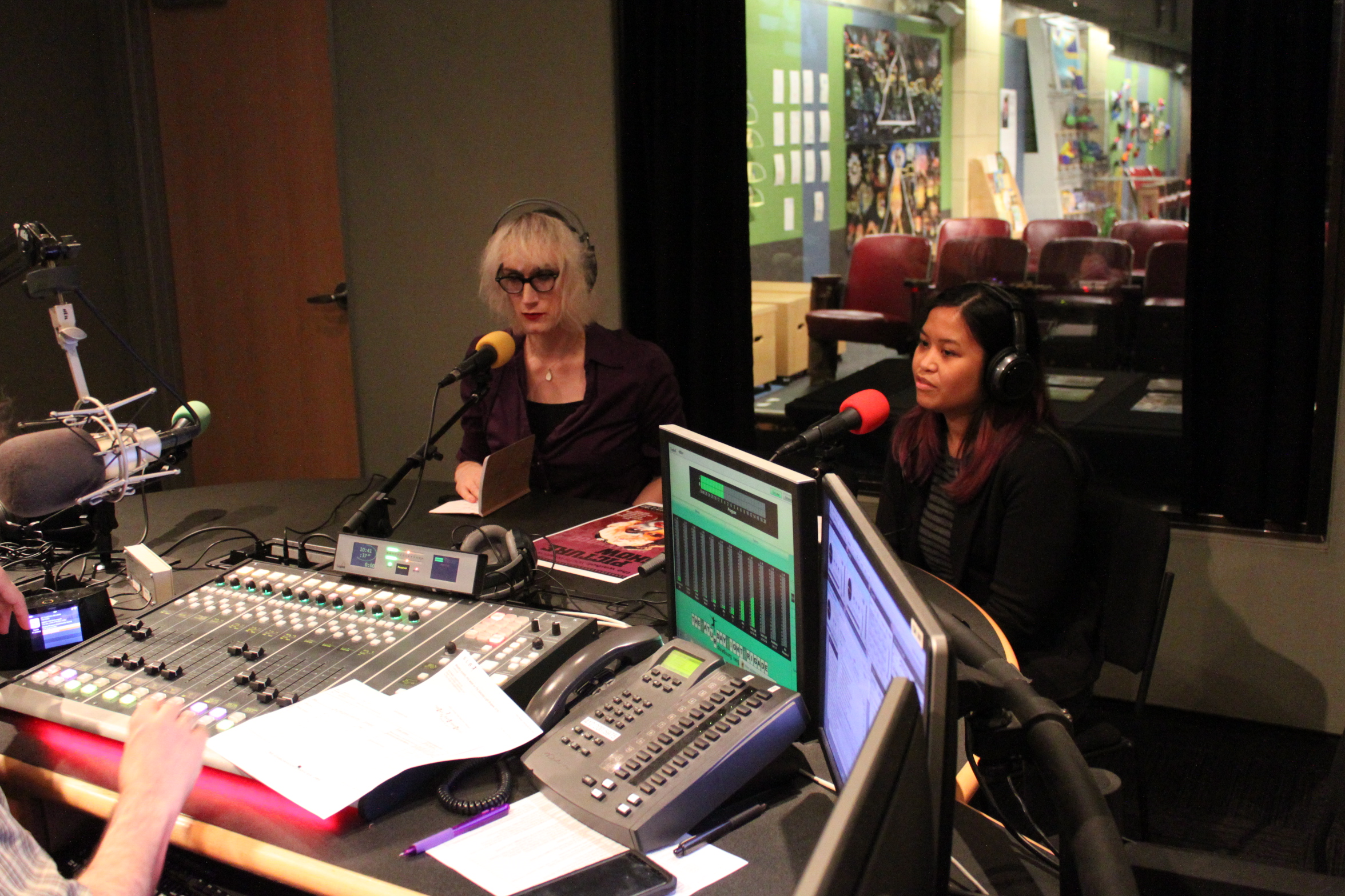 An adult and a teen both wearing headphones sitting behind microphones in a radio studio
