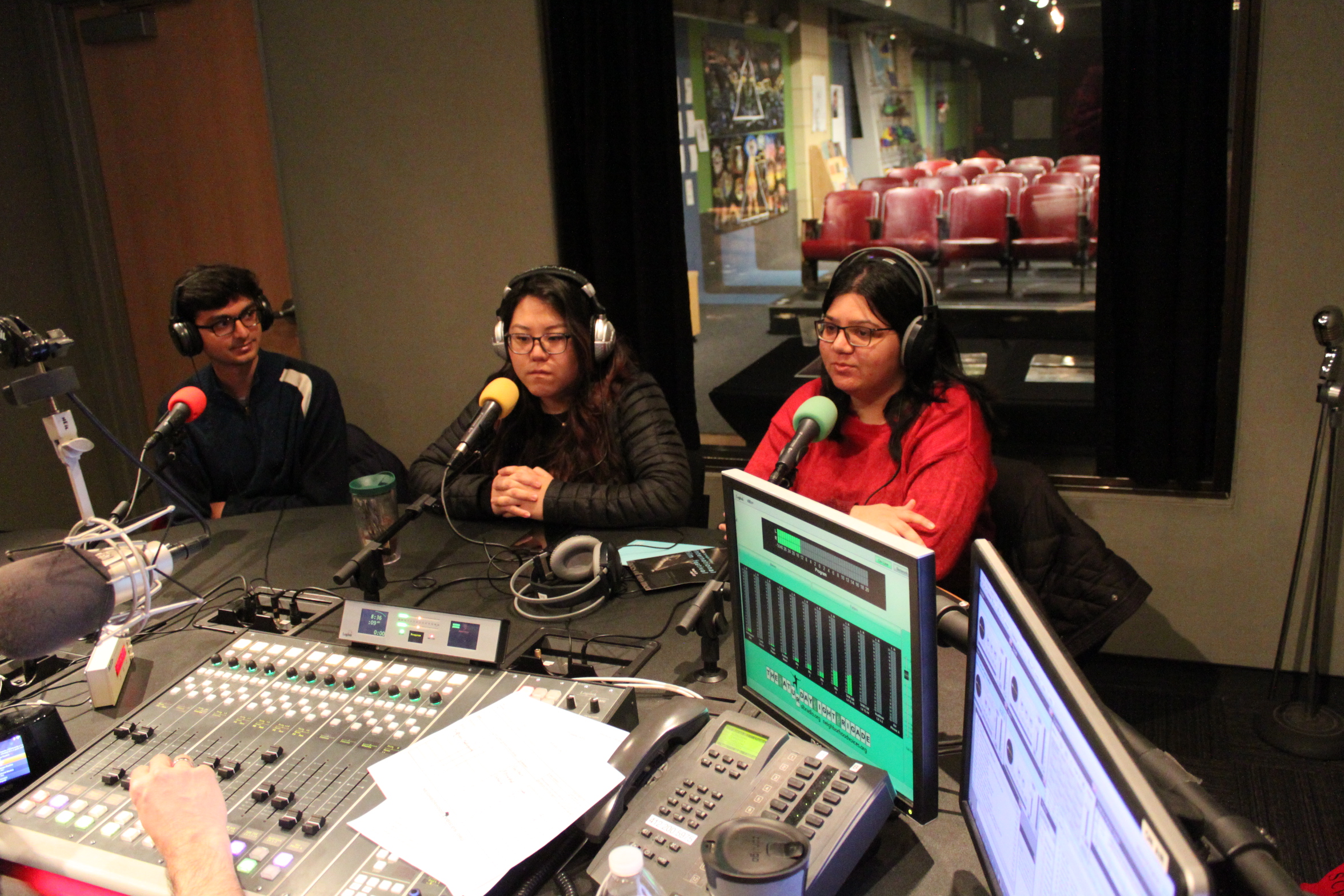 Three teens wearing headphones sitting behind microphones in a radio studio