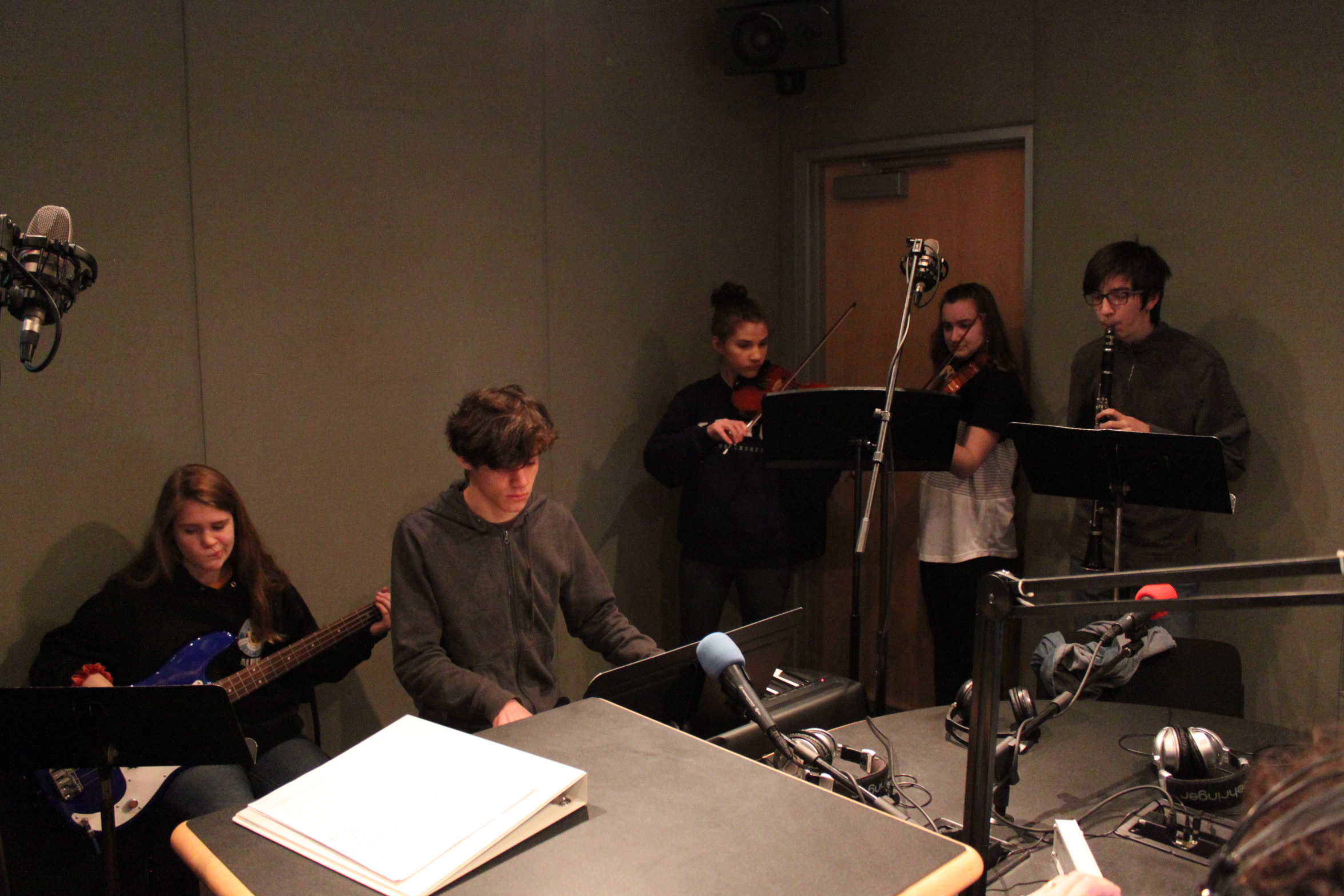 A teen playing bass guitar, a teen playing piano keyboard, two teens playing violin, and a teen playing clarinet, all looking at music stands in a radio studio