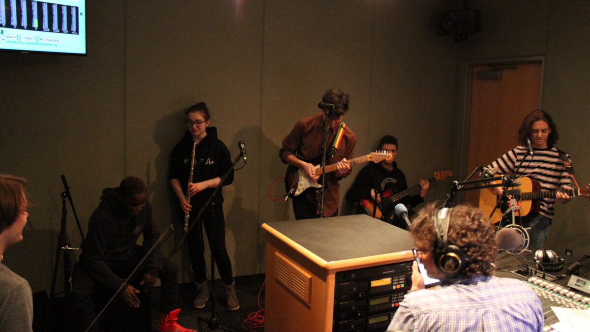 A teen playing electric violin, a teen playing djembe, a teen playing flute, a teen playing electric guitar, a teen playing bass, and a teen playing acoustic guitar as an adult wearing headphones sitting behind a microphone and a mixing board listens in a radio studio