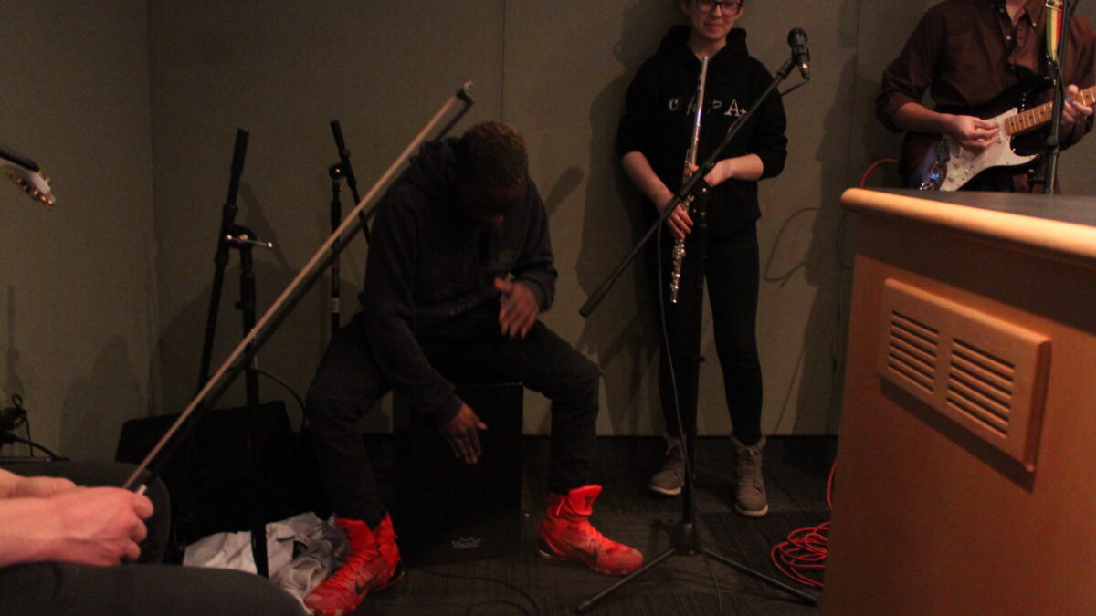 A teen playing djembe, a teen holding a flute, and a teen playing electric guitar in a radio studio