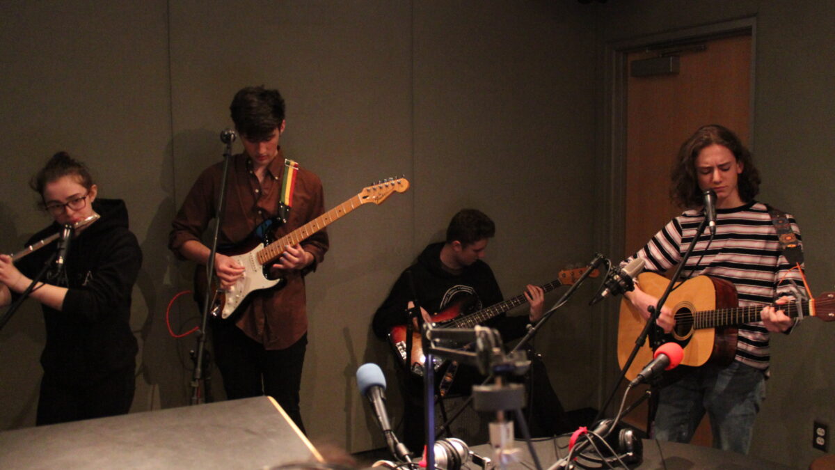 A teen playing flute, a teen playing electric guitar, a teen playing bass, and a teen playing acoustic guitar in a radio studio