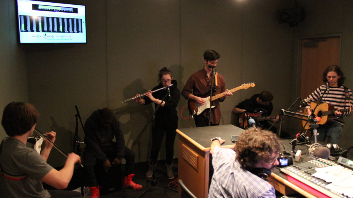 A teen playing electric violin, a teen playing djembe, a teen playing flute, a teen playing electric guitar, a teen playing bass, and a teen playing acoustic guitar in a radio studio as an adult wearing headphones listens
