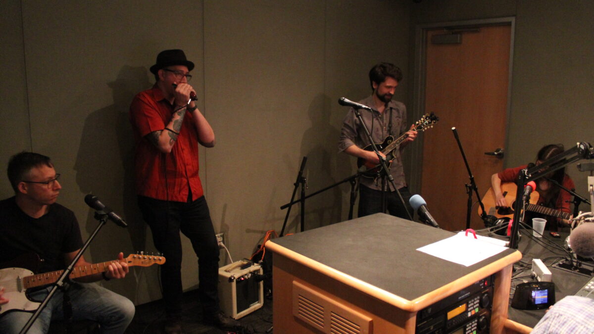 An adult playing electric guitar, an adult playing a harmonica, an adult playing mandolin, and an adult playing acoustic guitar in a radio studio