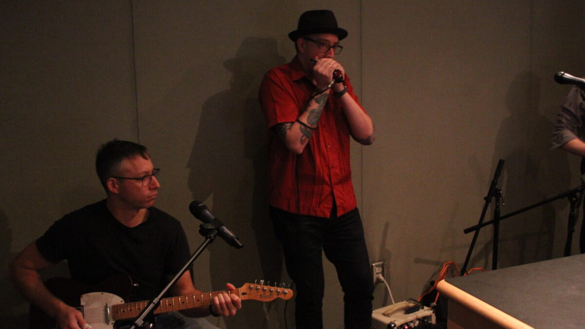 An adult playing guitar and an adult playing harmonica in a radio studio
