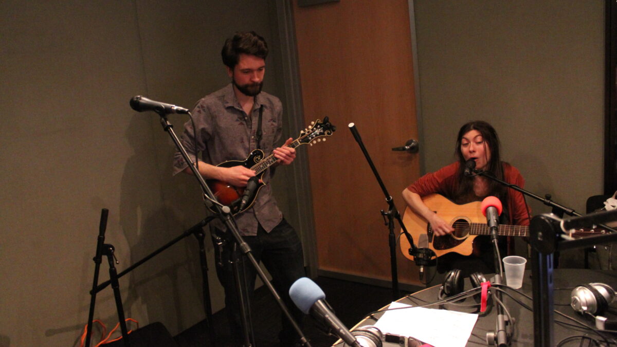 An adult playing a mandolin and an adult playing acoustic guitar in a radio studio