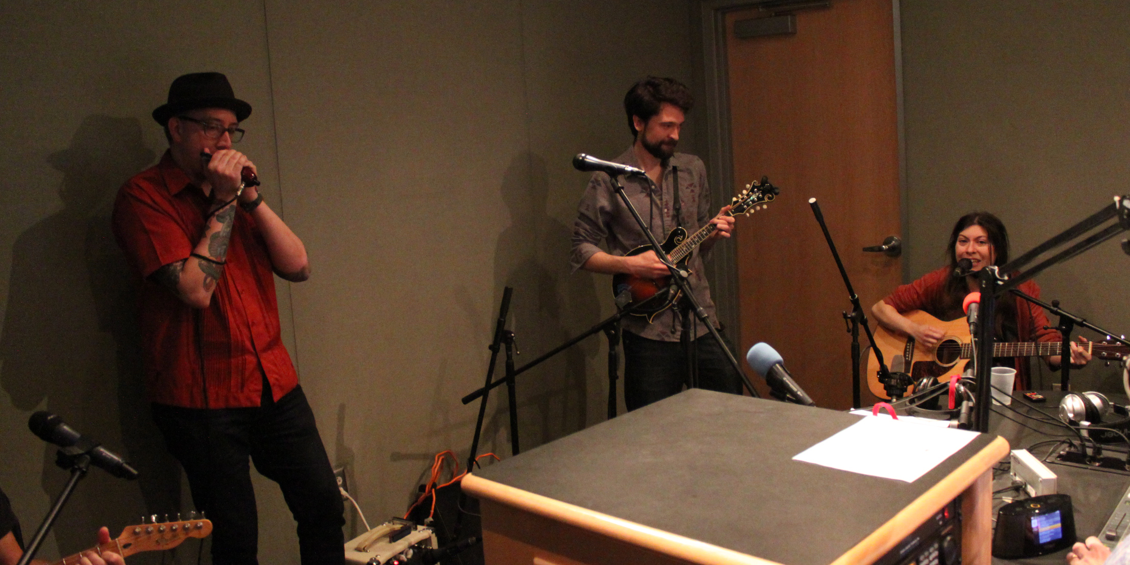 An adult playing harmonica, an adult playing ukulele, and an adult playing acoustic guitar in a radio studio
