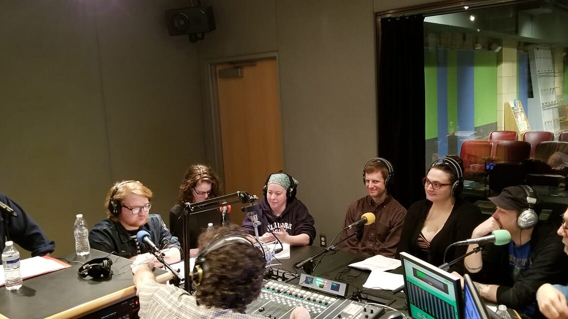 Eight adults wearing headphones sitting behind microphones being interviewed by an adult wearing headphones sitting behind a microphone and a mixing board in a radio studio