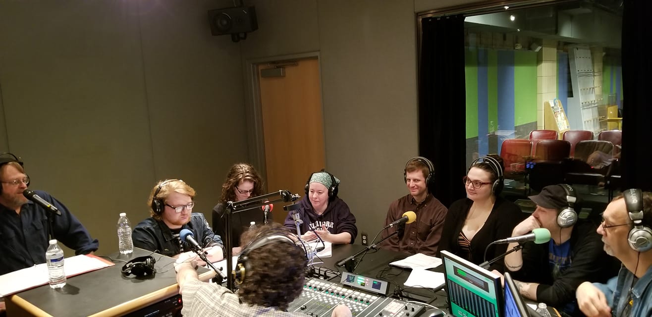 Eight adults wearing headphones sitting behind microphones being interviewed by an adult wearing headphones sitting behind a microphone and a mixing board in a radio studio