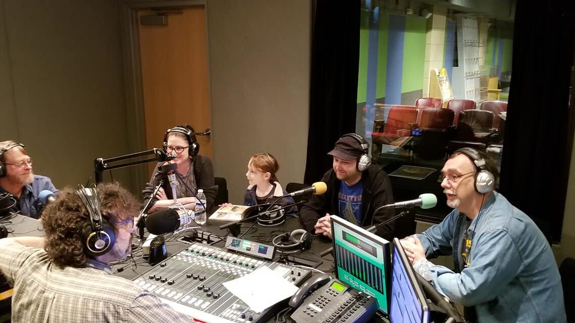 Four adults wearing headphones speaking into microphones and one child sitting behind a microphone being interviewed by an adult wearing headphones and speaking into a microphone sitting behind a mixing board in a radio studio