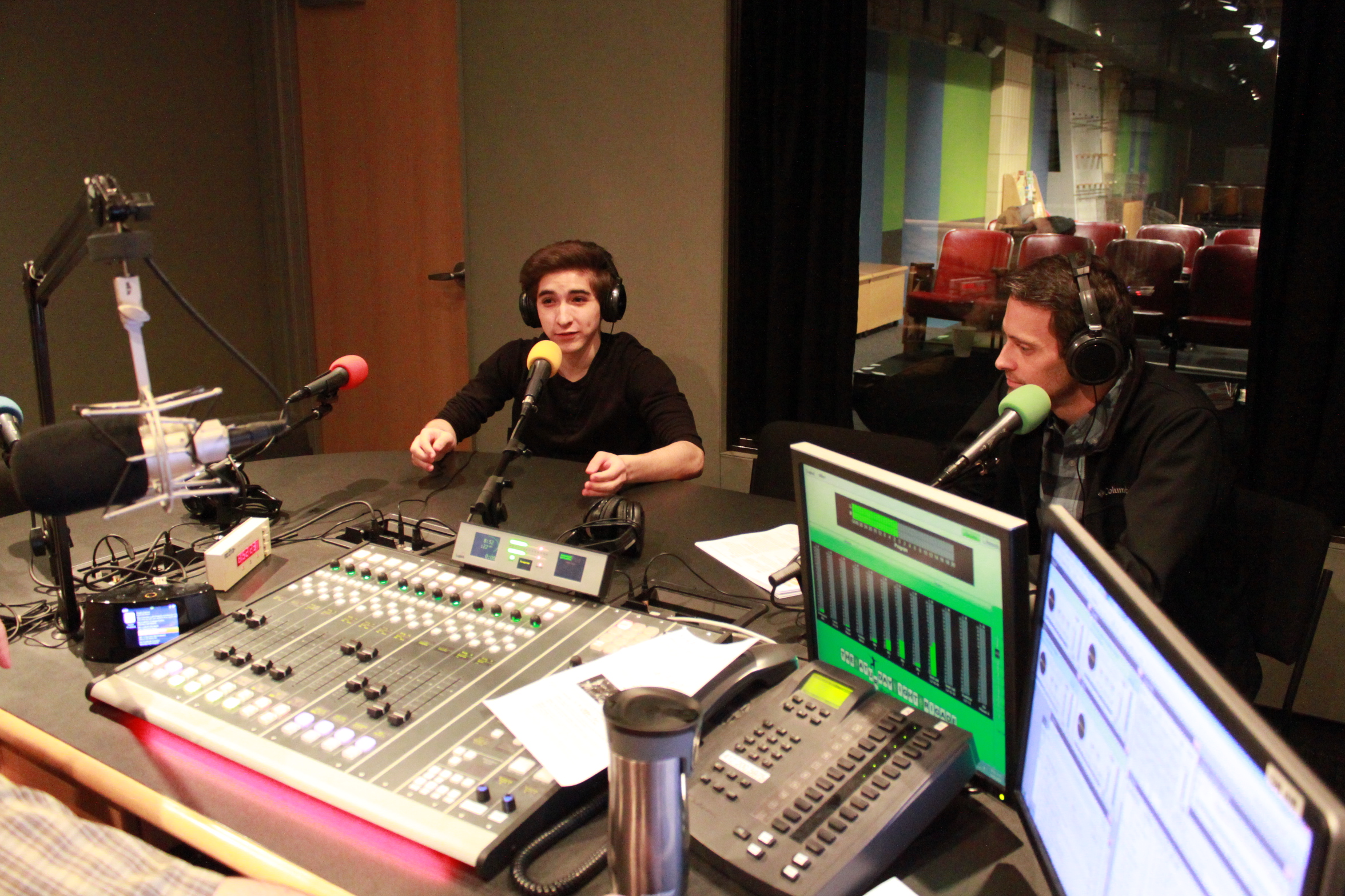 An adult wearing headphones sitting behind a microphone looking at a teen wearing headphones speaking into a microphone in a radio studio