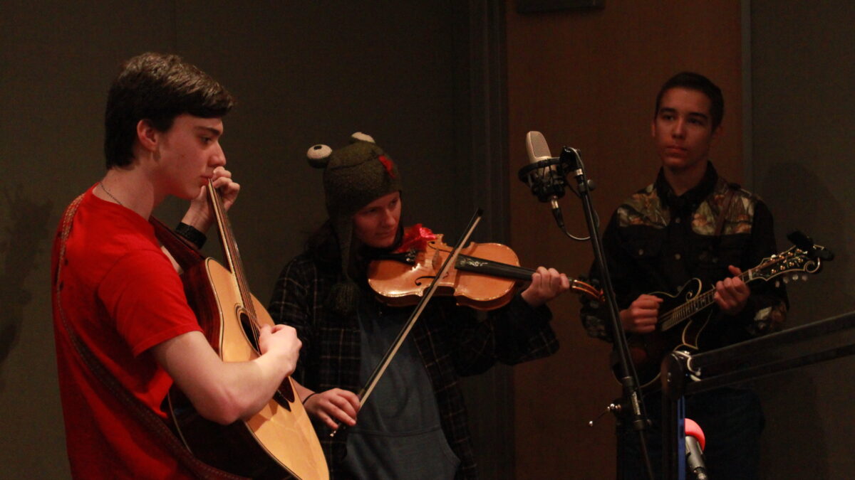 A teen playing guitar, a teen playing violin, and a teen playing mandolin in a radio studio