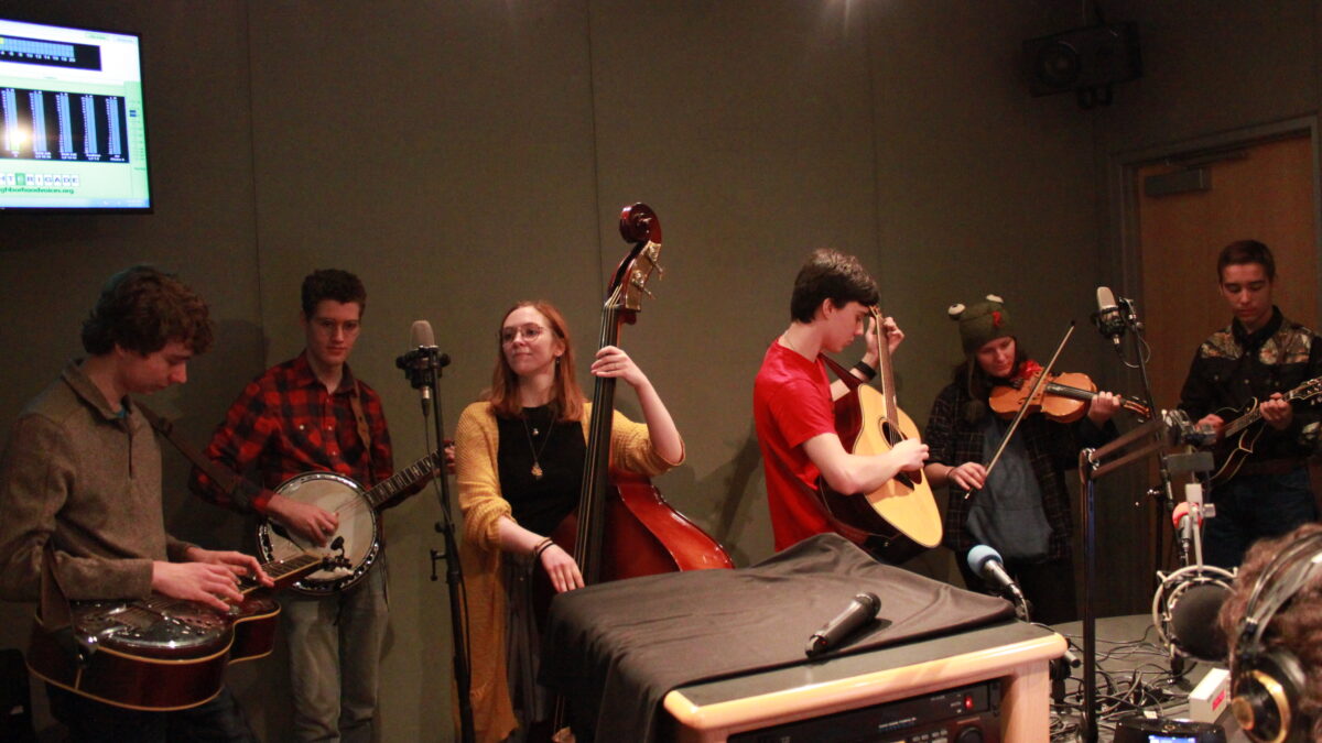 A teen playing dobro, a teen playing banjo, a teen playing upright bass, a teen playing guitar, a teen playing violin, and a teen playing mandolin in a radio studio