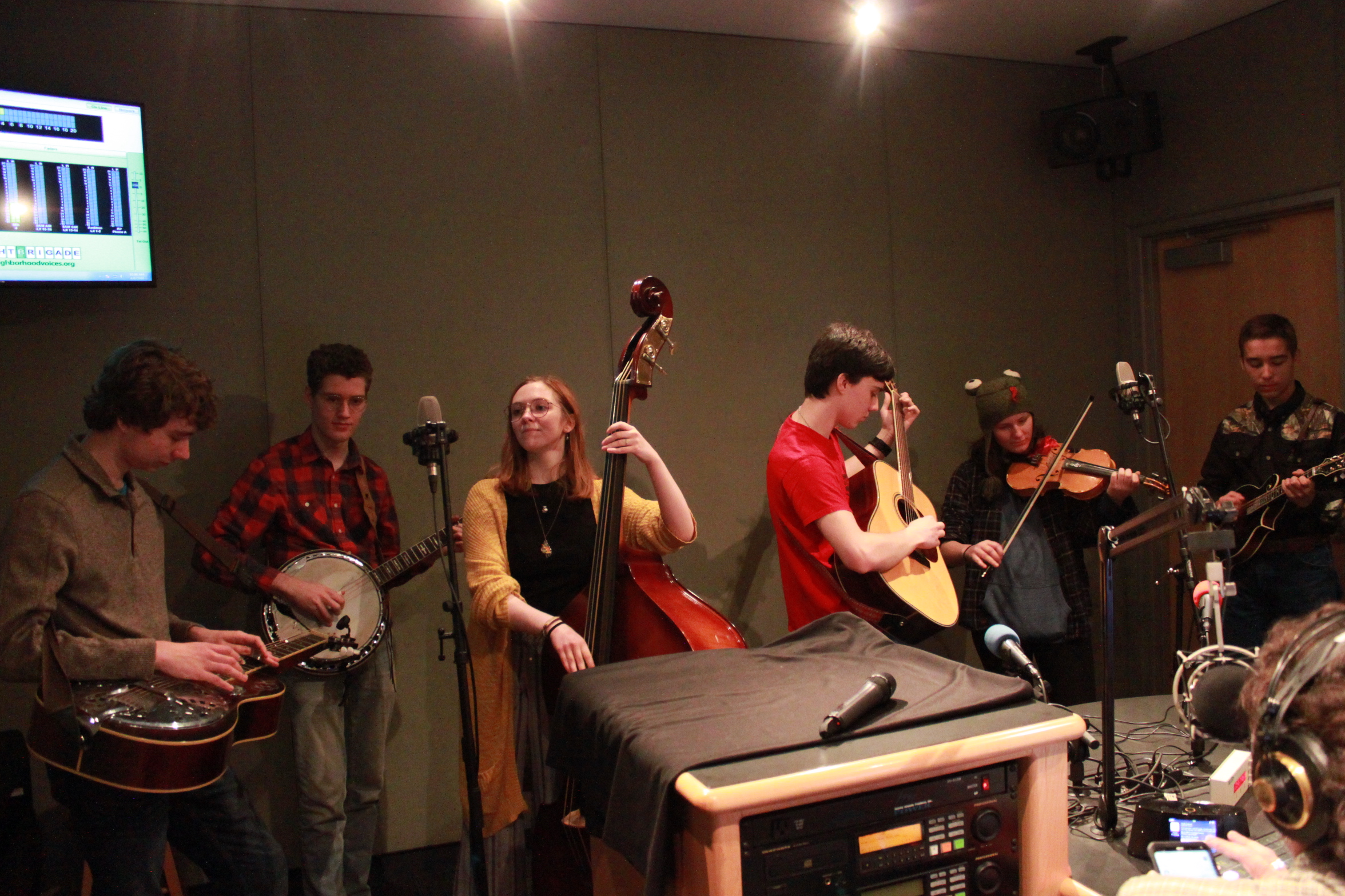 A teen playing dobro, a teen playing banjo, a teen playing upright bass, a teen playing guitar, a teen playing violin, and a teen playing mandolin in a radio studio