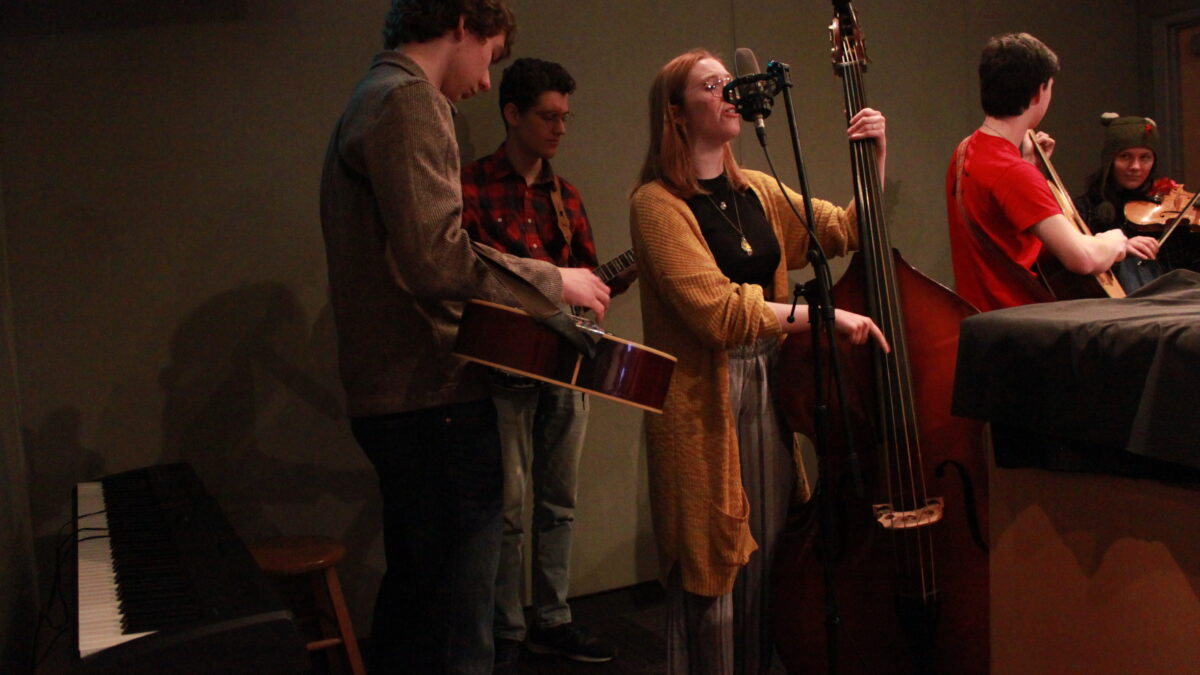 A teen playing dobro, a teen playing banjo, a teen playing upright bass and singing into a microphone, a teen playing guitar, and a teen playing violin in a radio studio