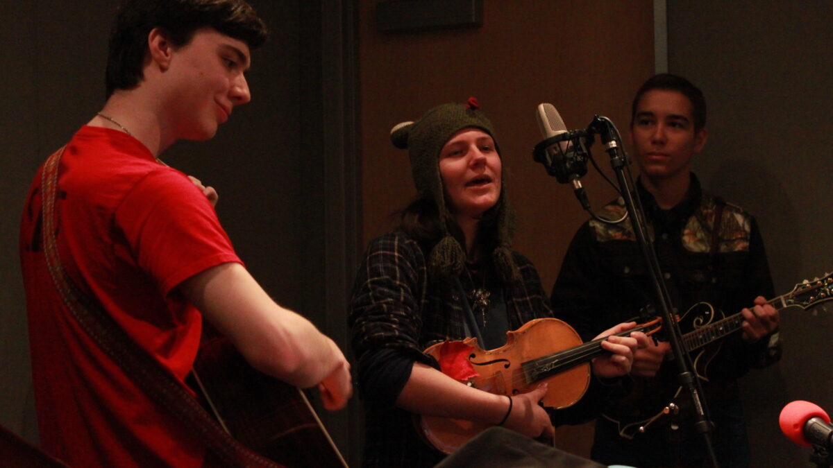 A teen playing guitar, a teen playing violin and singing into a microphone, and a teen playing mandolin in a radio studio