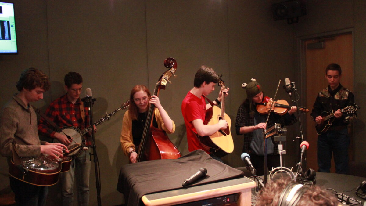 A teen playing dobro, a teen playing banjo, a teen playing upright bass, a teen playing guitar, a teen playing violin, and a teen playing mandolin in a radio studio