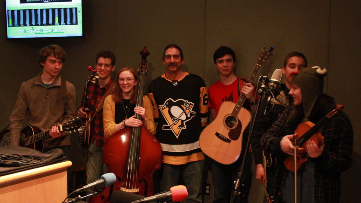 A teen holding a dobro, a teen holding a banjo, a teen holding an upright bass, an adult with their hands behind their back, a teens holding a guitar, a teen holding a mandolin, and a teen holding a violin in a radio studio
