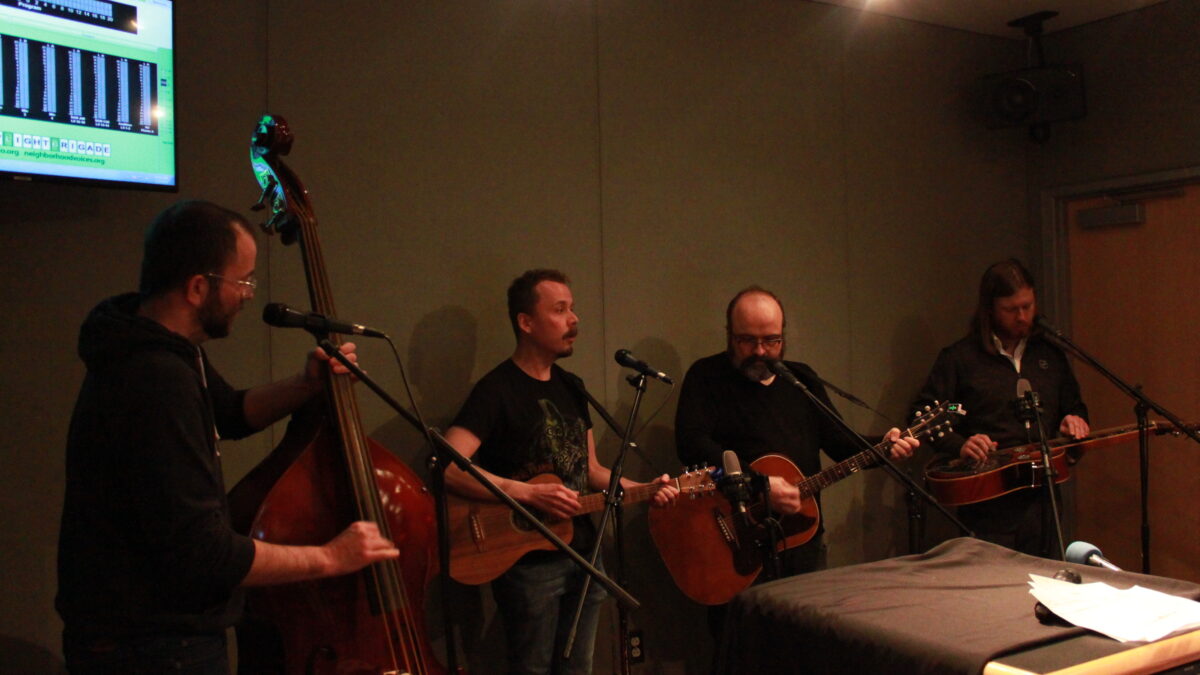 An adult playing upright bass, an adult playing bass ukulele, an adult playing guitar, and an adult playing dobro in a radio studio