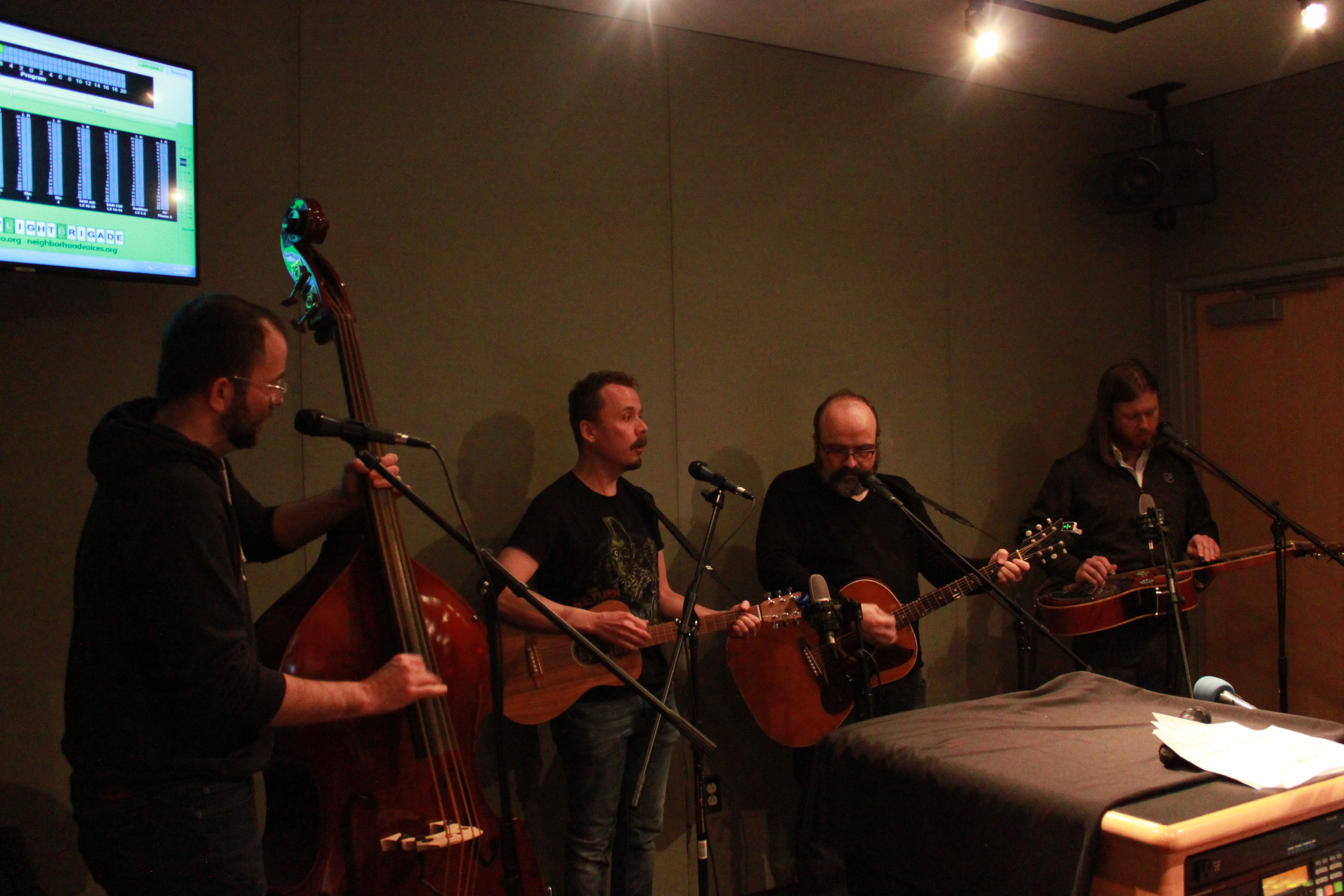 An adult playing upright bass, an adult playing bass ukulele, an adult playing guitar, and an adult playing dobro in a radio studio