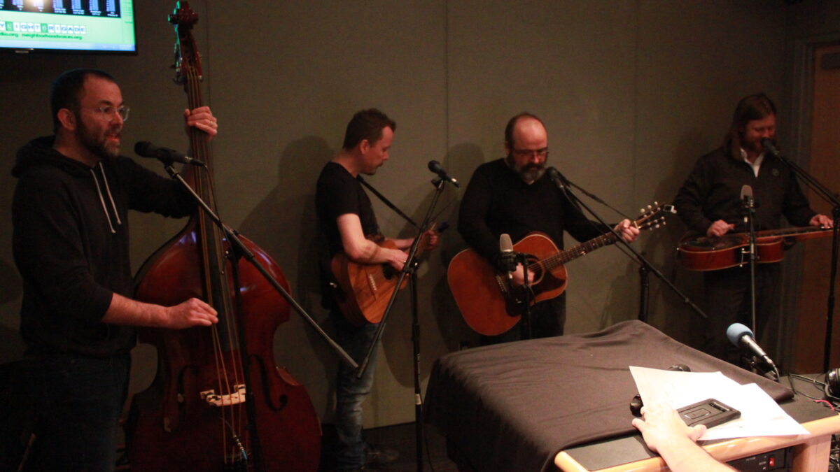 An adult playing upright bass, two adults playing guitar, and an adult playing dobro and singing into a microphone in a radio studio