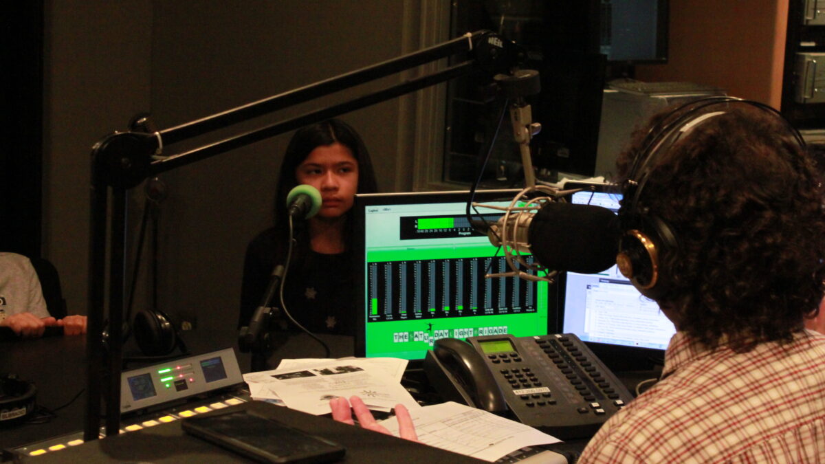 A youth sitting behind a microphone being interviewed by an adult wearing headphones sitting behind a microphone and a mixing board in a radio studio