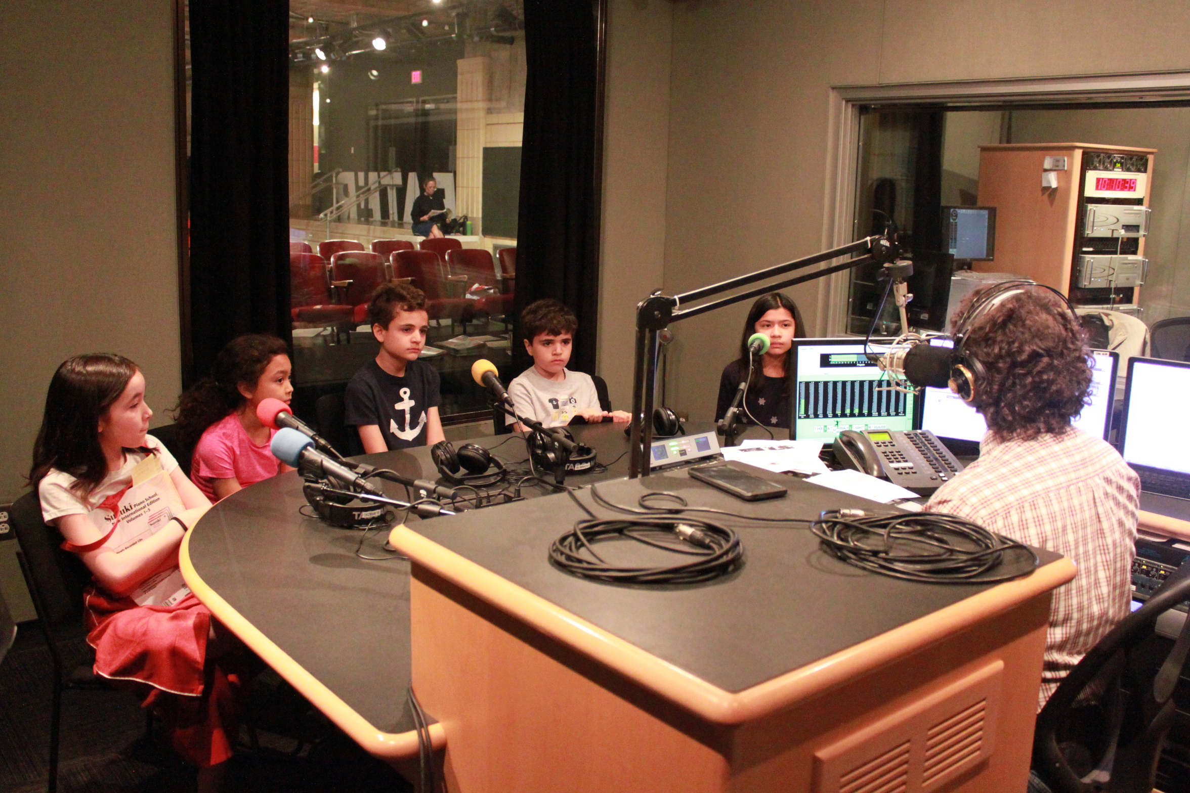 Five youth sitting behind microphones being interviewed by an adult wearing headphones sitting behind a microphone and a mixing board in a radio studio