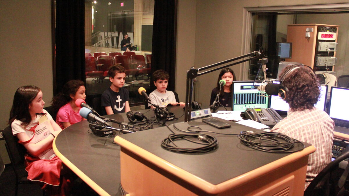 Five youth sitting behind microphones being interviewed by an adult wearing headphones sitting behind a microphone and a mixing board in a radio studio