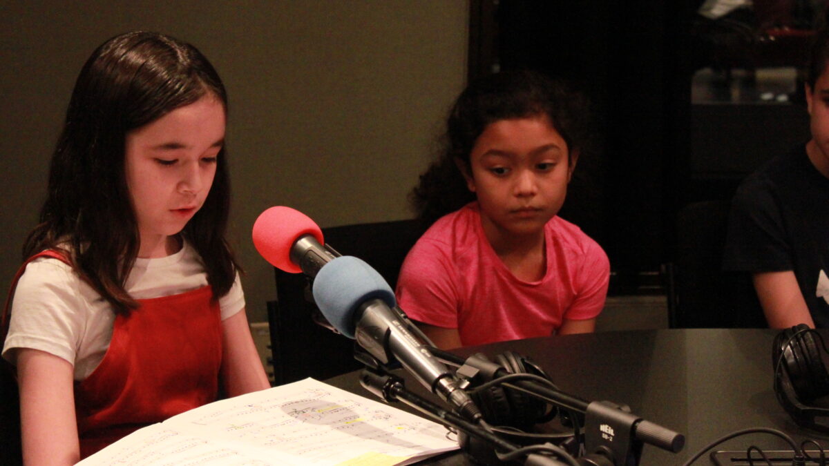 Three youth sitting behind microphones in a radio studio, one youth looking down at sheet music on a table