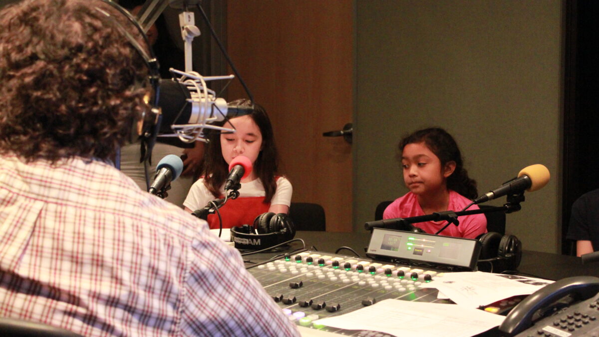 Two youth speaking into microphones being interviewed by an adult wearing headphones and sitting behind a microphone and a mixing board in a radio studio