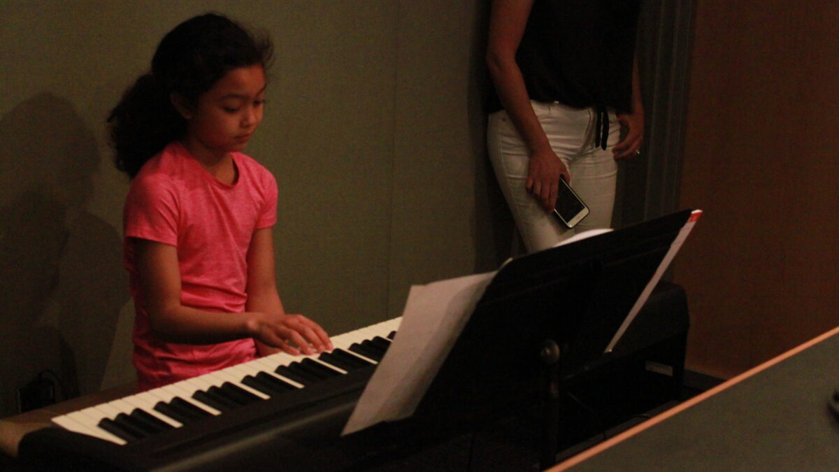A child playing a piano keyboard looking at sheet music on a music stand and an individual holding a smartphone in a radio studio
