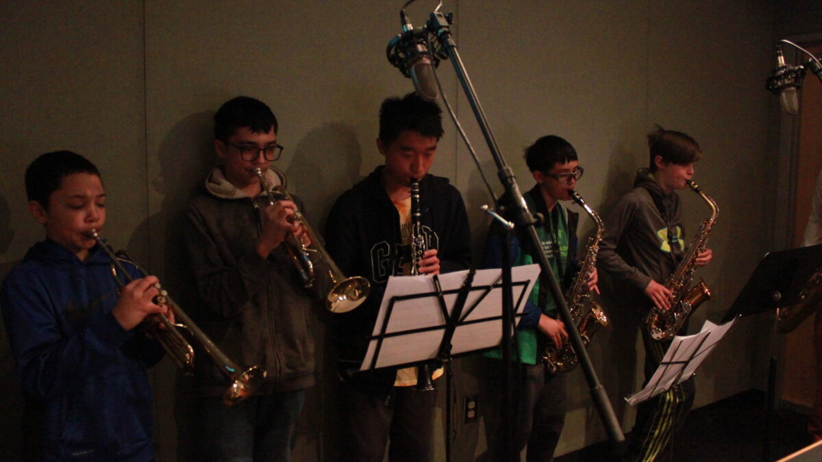 Two youth playing trumpet, one youth playing clarinet, and two youth playing alto saxophone, all looking at sheet music on music stands in a radio studio