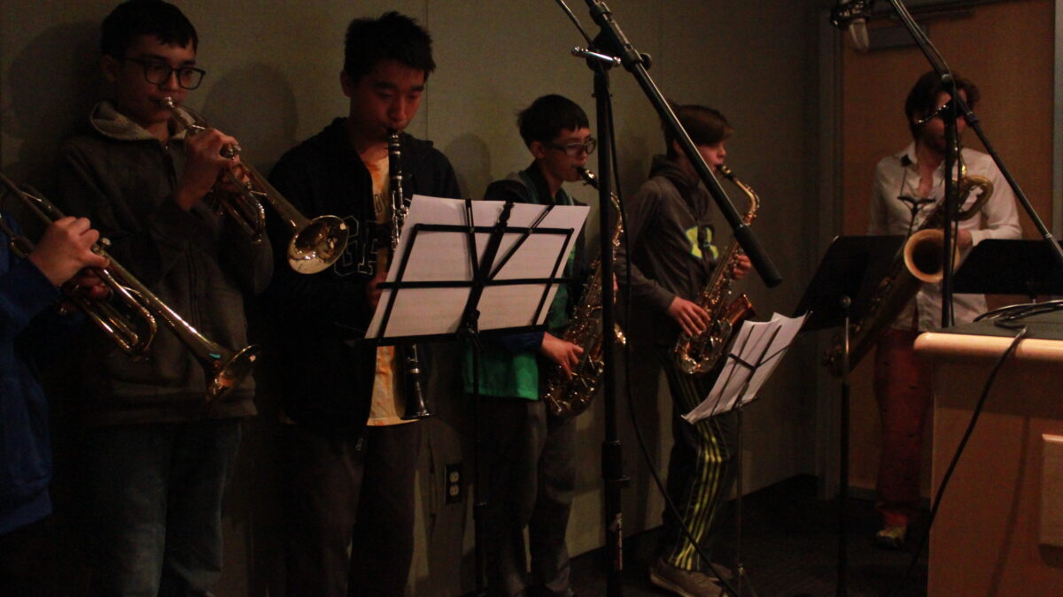 Two youth playing trumpet, one youth playing clarinet, two youth playing alto saxophone, and one adult playing baritone saxophone, all looking at sheet music on music stands in a radio studio