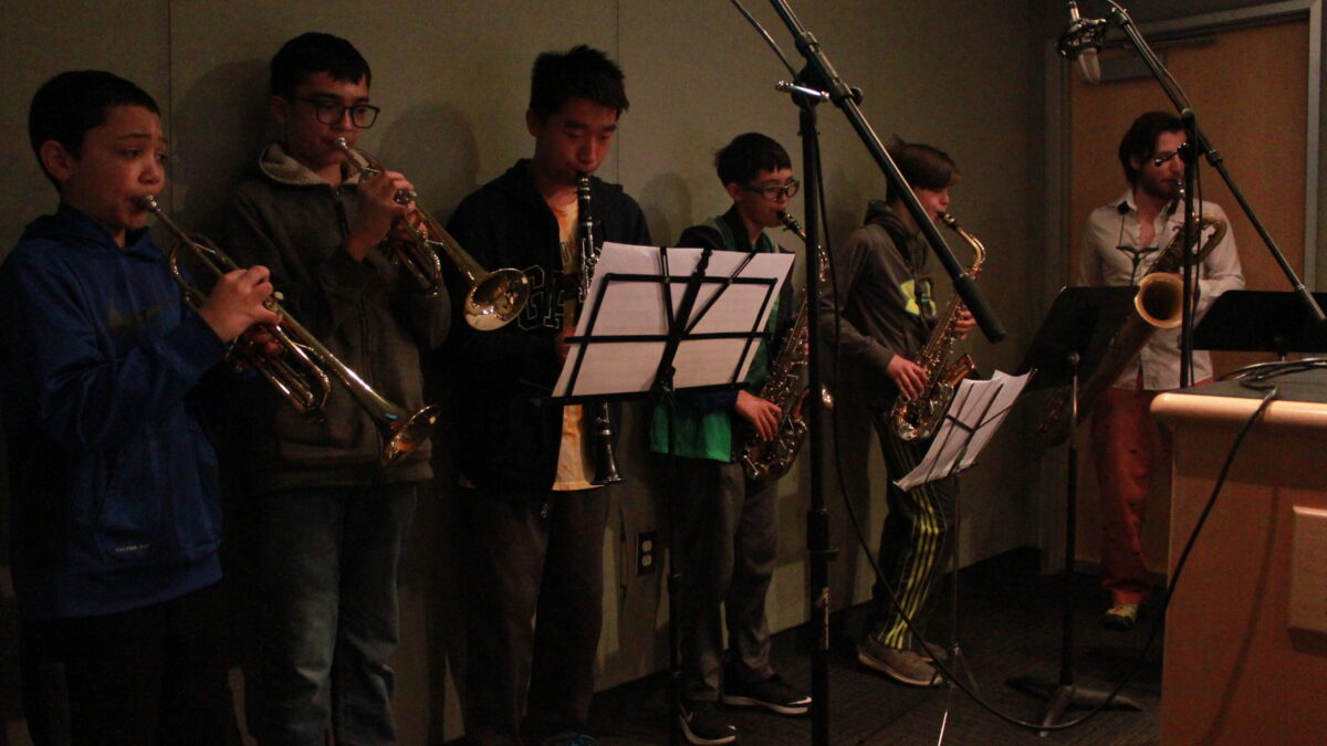 Two youth playing trumpet, one youth playing clarinet, two youth playing alto saxophone, and one adult playing baritone saxophone, all looking at sheet music on music stands in a radio studio