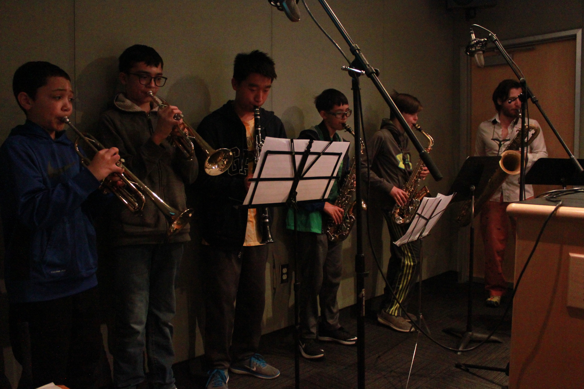 Two youth playing trumpet, one youth playing clarinet, two youth playing alto saxophone, and one adult playing baritone saxophone, all looking at sheet music on music stands in a radio studio