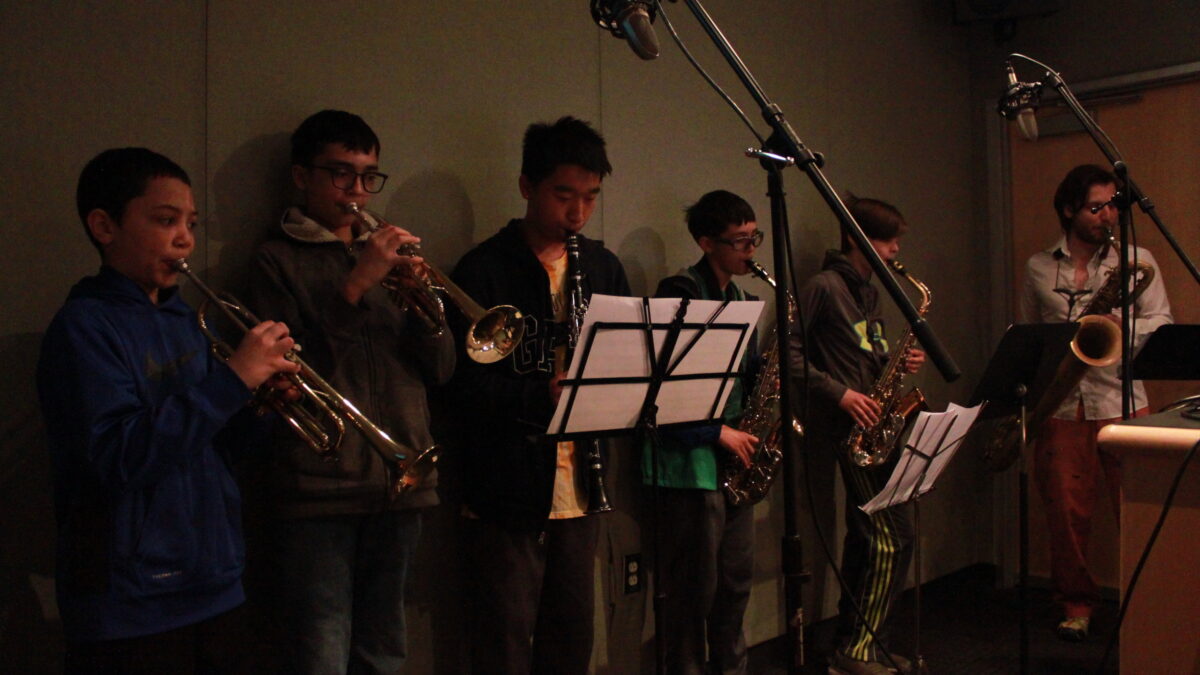 Two youth playing trumpet, one youth playing clarinet, two youth playing alto saxophone, and an adult playing baritone saxophone, all looking at sheet music on music stands in a radio studio