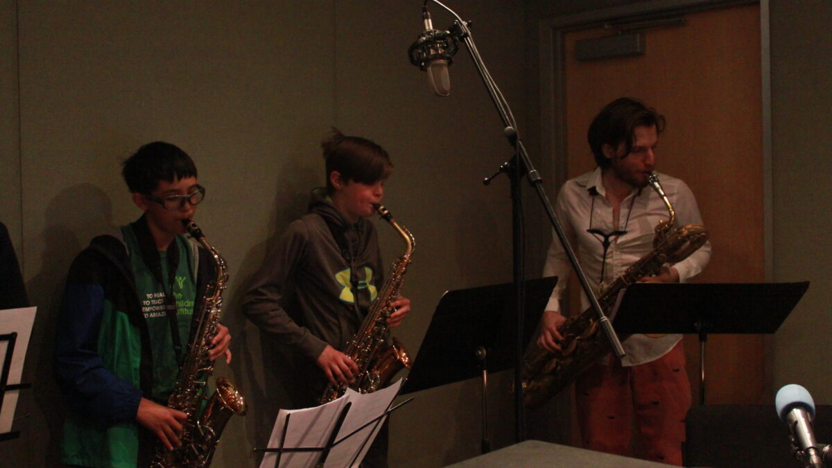 Two youth playing alto saxophone and an adult playing baritone saxophone, all looking at sheet music on music stands in a radio studio