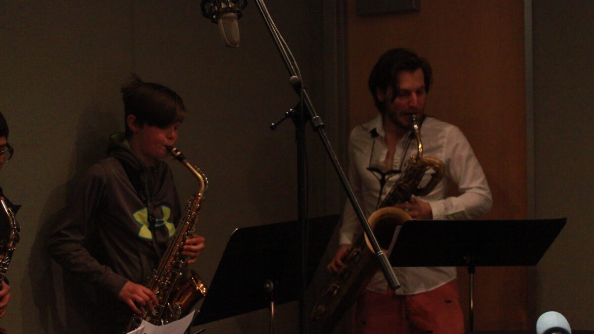 Two youth playing alto saxophone and an adult playing baritone saxophone, all looking at sheet music on music stands in a radio studio