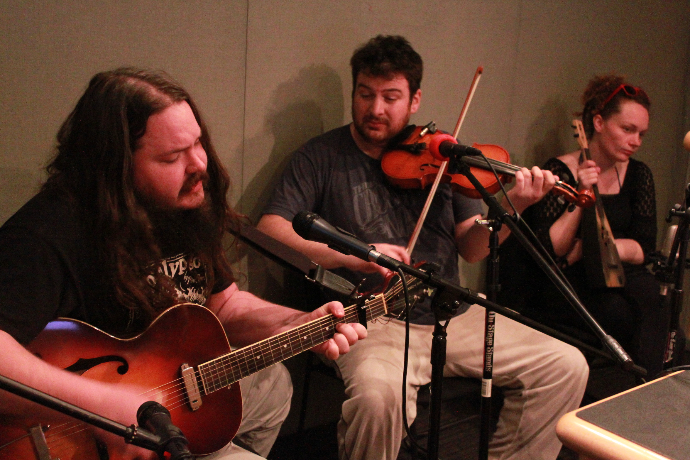 An adult playing guitar, an adult playing violin, and an adult holding a ukulele in a radio studio