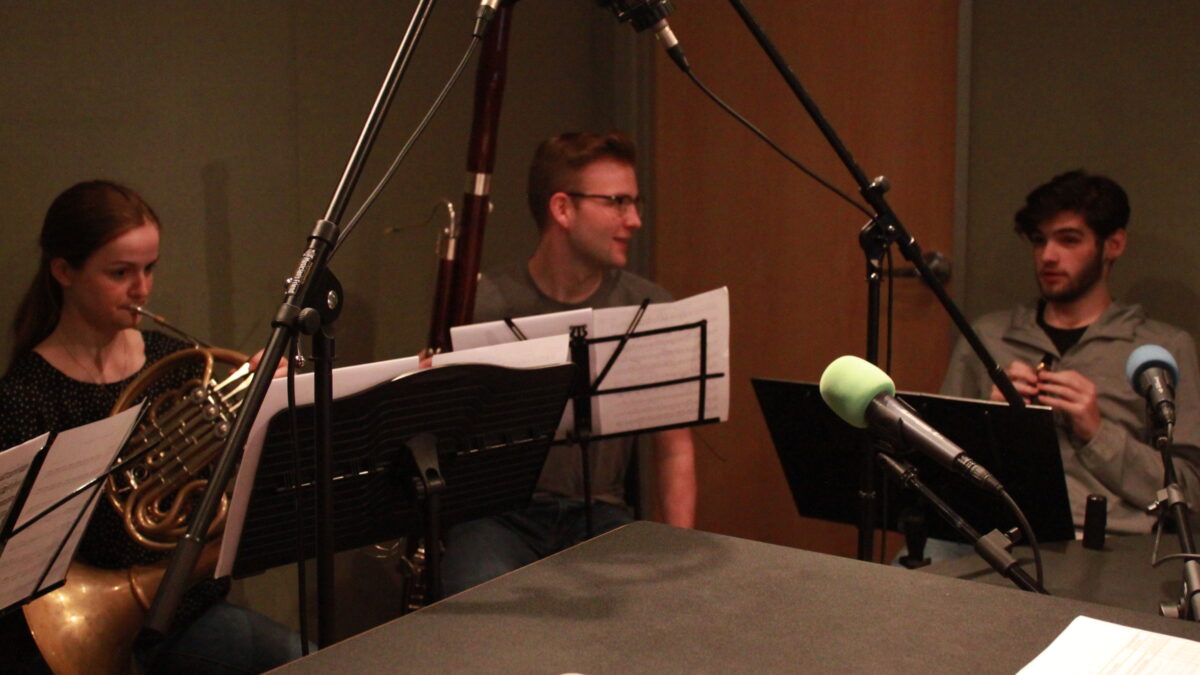 A teen playing french horn, a teen holding a bassoon, and a teen holding a clarinet, all looking at sheet music on music stands in a radio studio