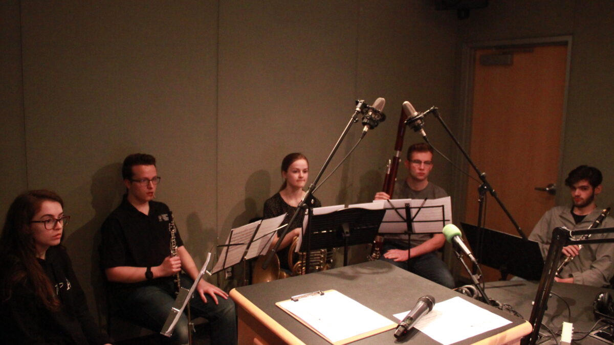 A teen holding a flute, a teen holding an oboe, a teen holding a french horn, a teen holding a bassoon, and a teen holding a clarinet, all sitting behind sheet music on music stands in a radio studio
