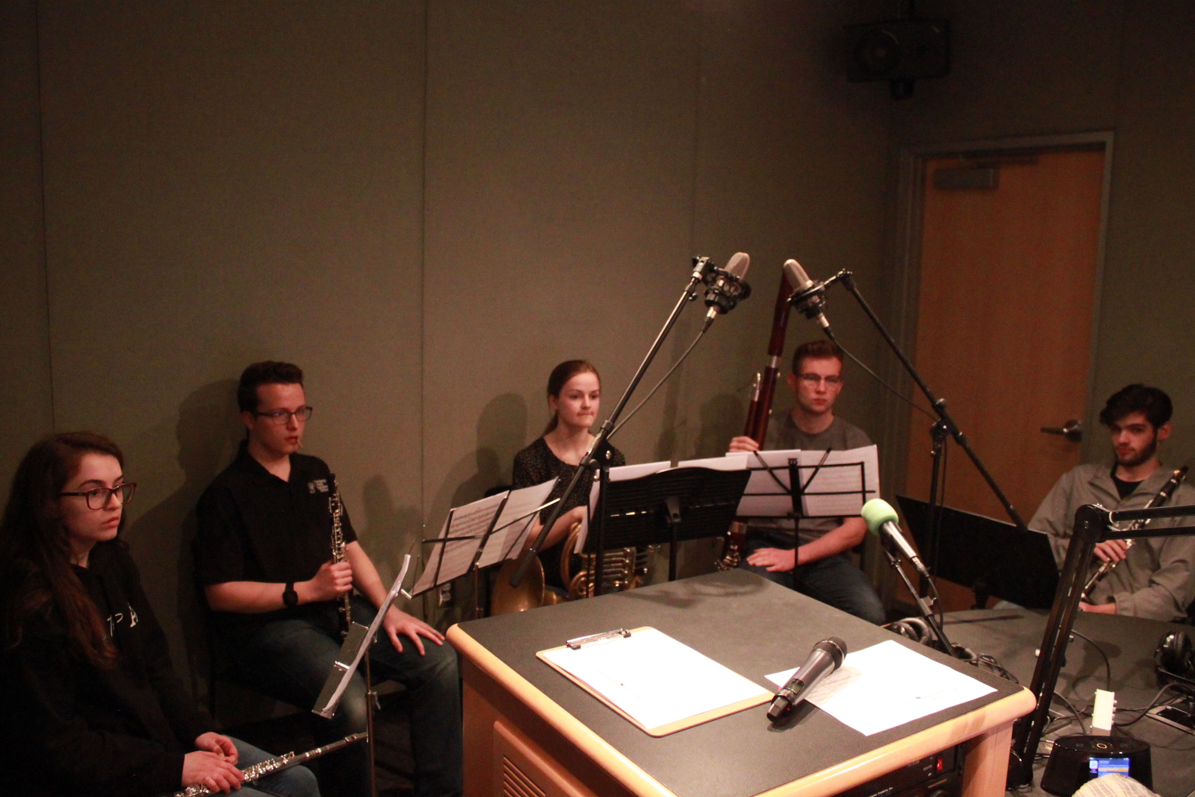 A teen holding a flute, a teen holding an oboe, a teen holding a french horn, a teen holding a bassoon, and a teen holding a clarinet, all sitting behind sheet music on music stands in a radio studio