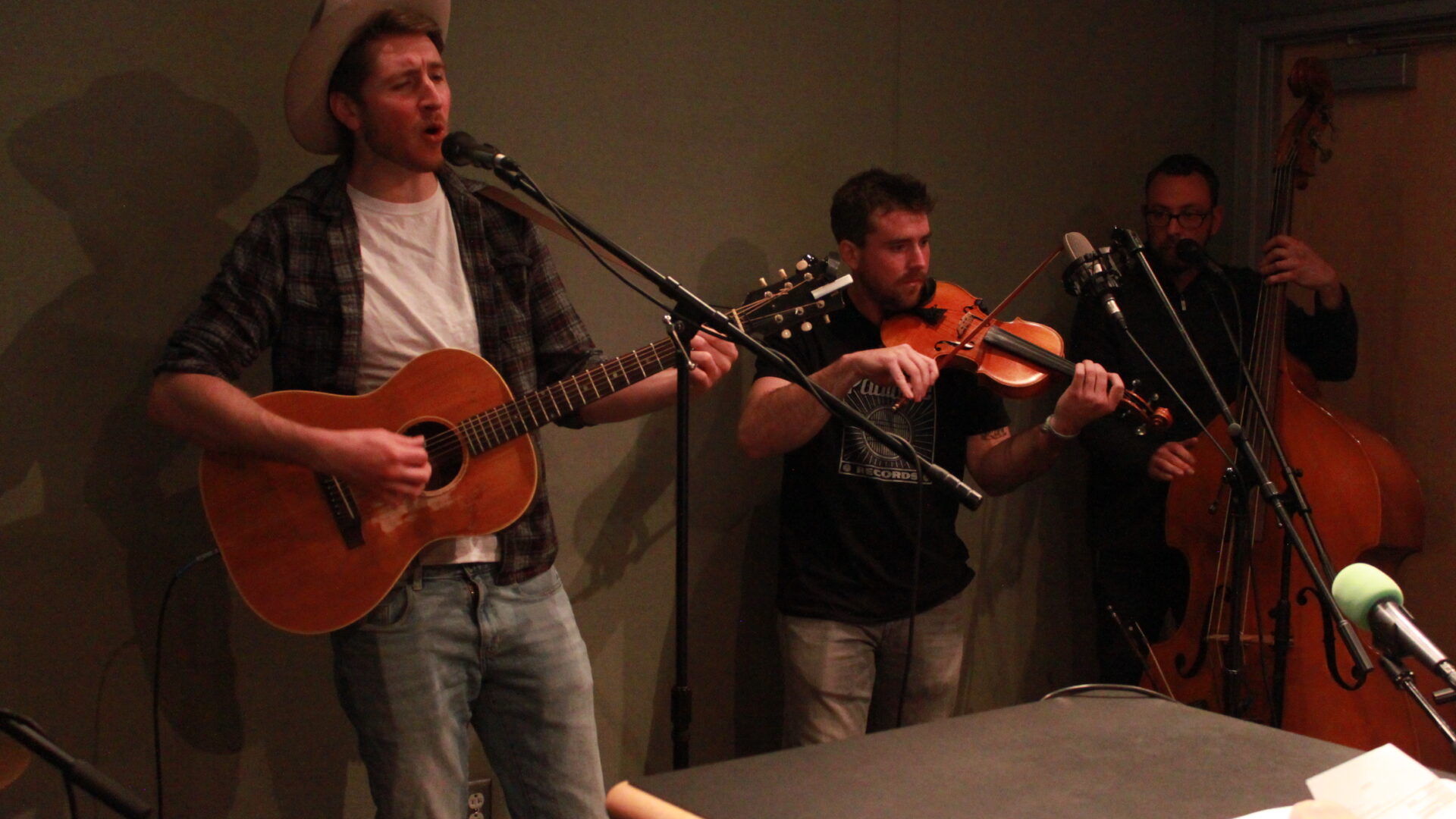 An adult playing guitar and singing into a microphone, an adult playing violin, and an adult playing upright bass in a radio studio