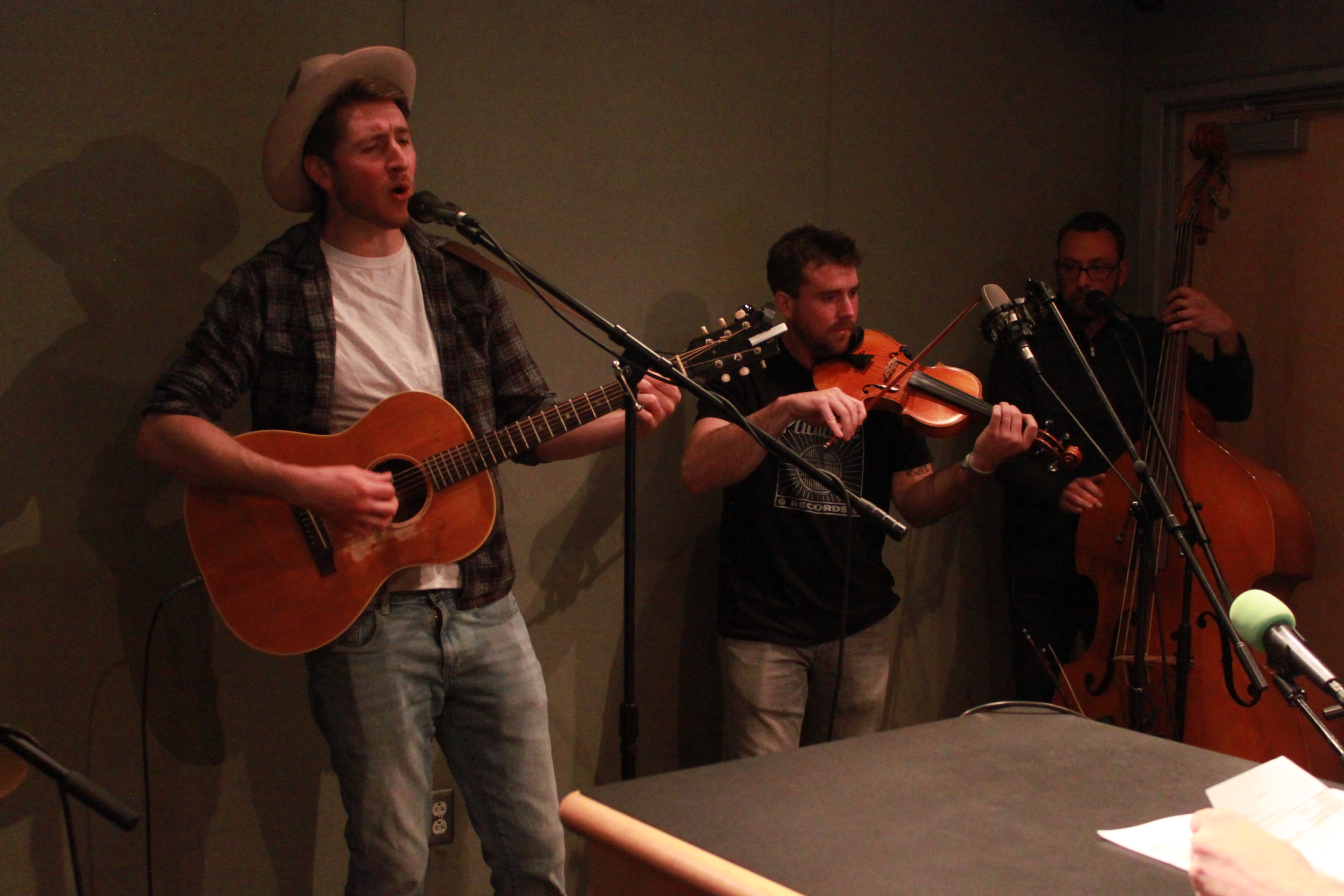 An adult playing guitar and singing into a microphone, an adult playing violin, and an adult playing upright bass in a radio studio