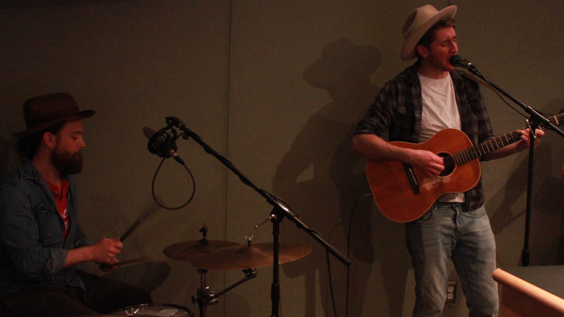 A adult playing drum set and an adult playing guitar and singing into a microphone in a radio studio