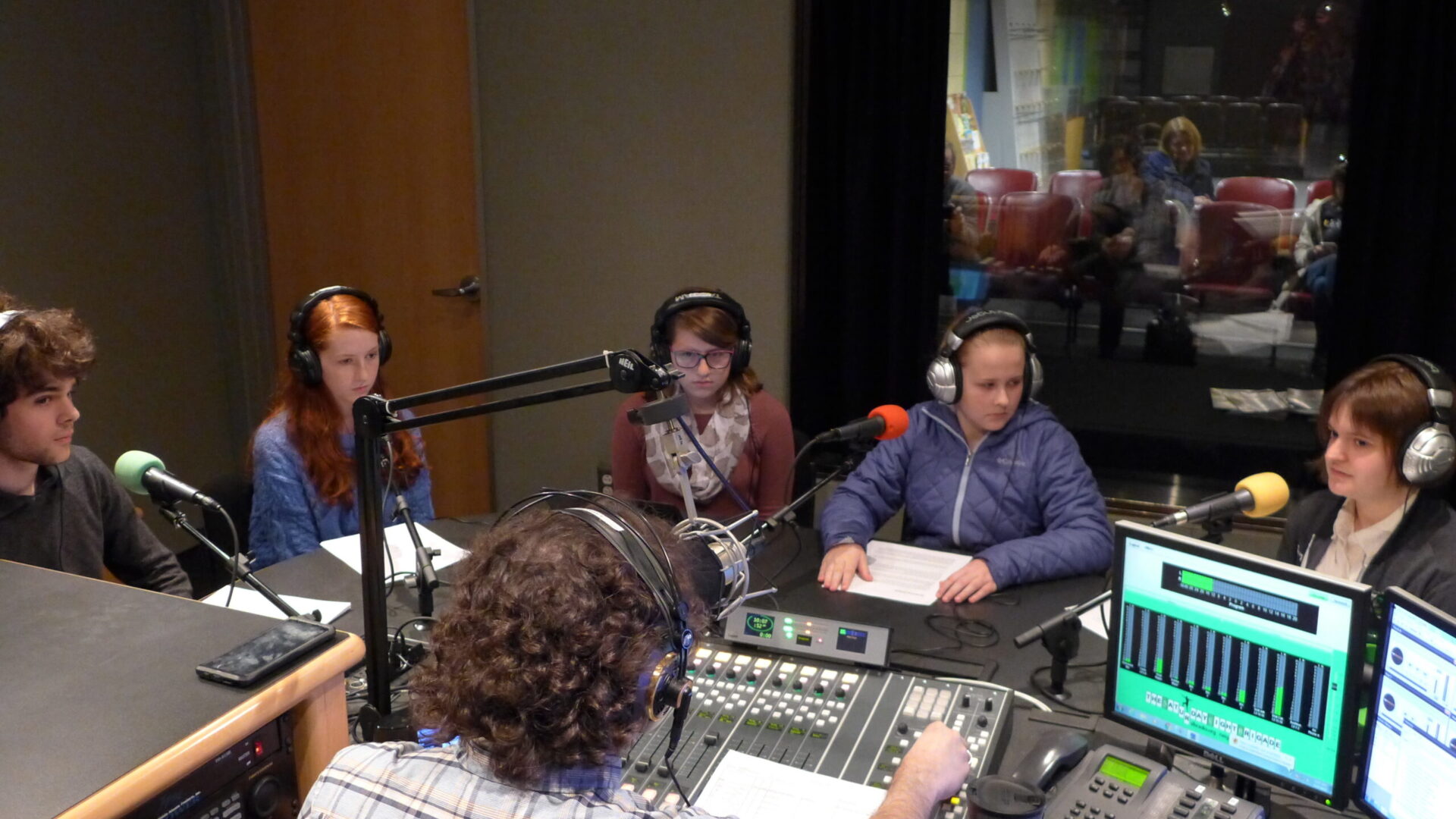 Five teens wearing headphones sitting behind microphones being interviewed by an adult wearing headphones sitting behind a microphone and a mixing board in a radio studio