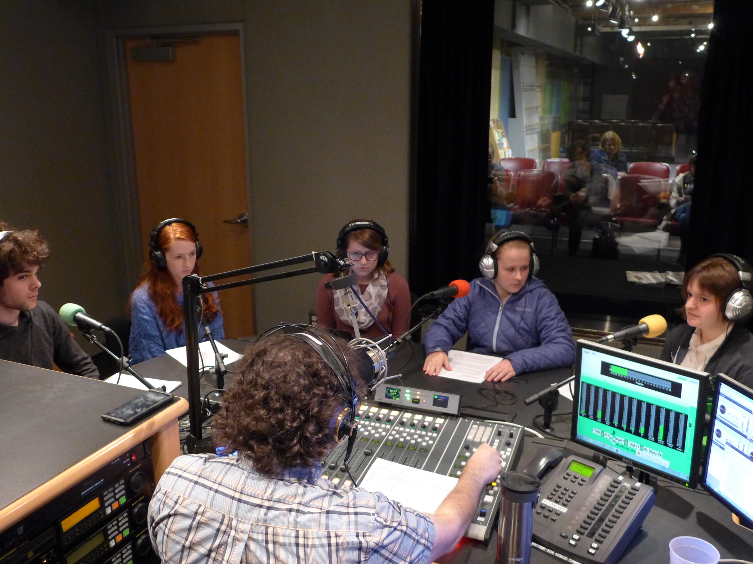 Five teens wearing headphones sitting behind microphones being interviewed by an adult wearing headphones sitting behind a microphone and a mixing board in a radio studio